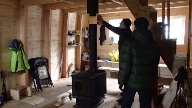 Workers are busy installing the black smoke stack of the wood stove. Sunlight shines in through the windows and gear is piled on temporary shelves in the background.