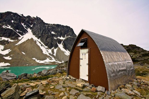 The Gothic Arch Hut front entrance with its brown front end-wall and bright white door sits next to the turquoise water of the glacial lake. A dark peak dotted with snow is visible in the background.