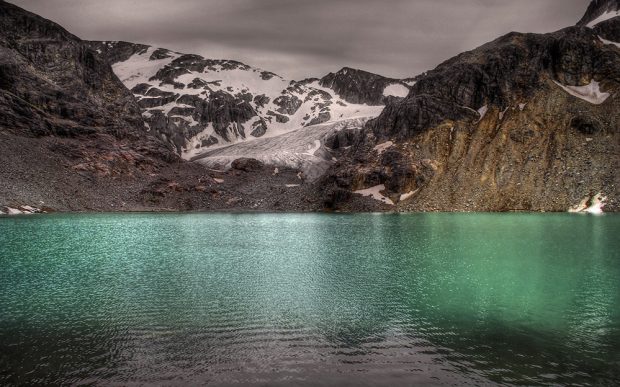 The turquoise water reflects against a glimmer of sunlight. Behind lies the dark brown rocky banks and the tip of the glacier rises up towards the bare rocky peak and the grey clouds.