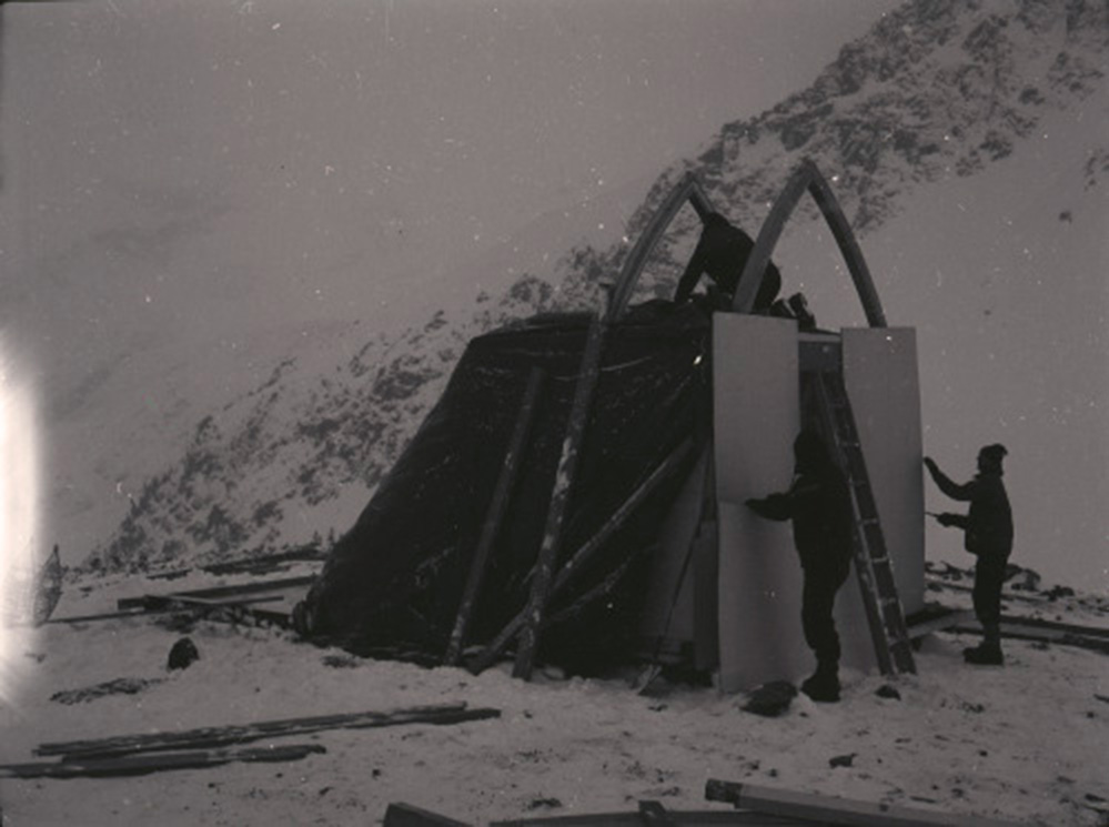 Snow covers the ground and the slope and three members of the construction crew attempt to assembly the Hut.