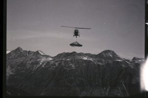 À l’arrière-plan se trouve une chaîne de montagnes avec de petits amas de neige. Au premier plan, l’hélicoptère transporte un chargement suspendu à un câble sous le cockpit.