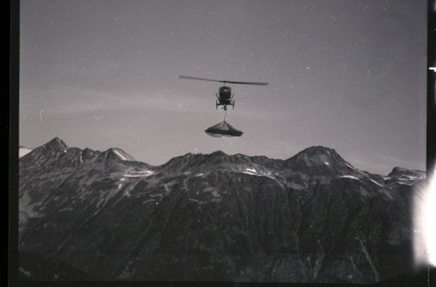 In the background, sits a range of mountains with small patches of snow. The flying helicopter is in the foreground carrying a load underneath the cockpit dangling from a cable.