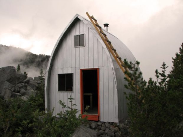 The Harrison Hut standing wrapped completely in aluminum siding following renovations in August 2014. Grey clouds swirl above in the overcast sky and the evergreen trees stand out against the shiny silver of the finished Hut.
