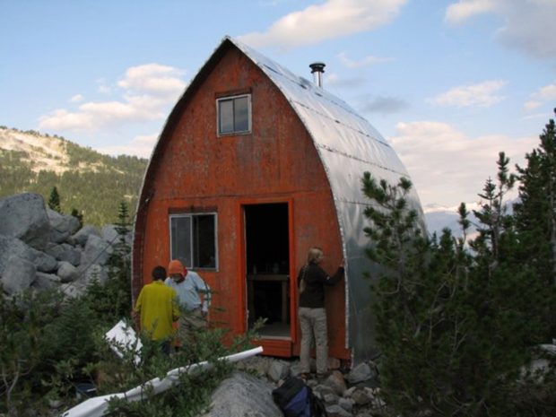 Two men stand in front of the weathered Hut and a woman inspects the wall near the open front door. The blue sky is dotted with white puffy clouds and sunshine can be seen reflecting off the evergreen trees in the distance.