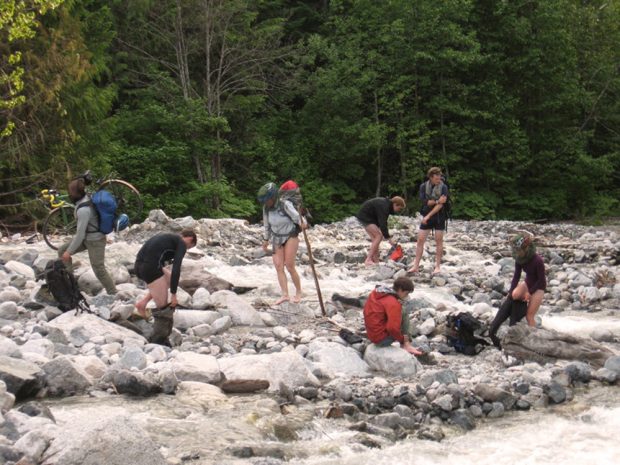 A group of UBC-VOC members use the flowing creek to get clean following work on the trail to the Harrison Hut. One of the members is carrying a bicycle and bags across the flowing creek.