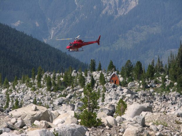 Bright red helicopter in the air above the Harrison Hut prior to renovations. The orange end-wall is visible in the boulder field surrounding it. Short green evergreen trees grow near the hut.