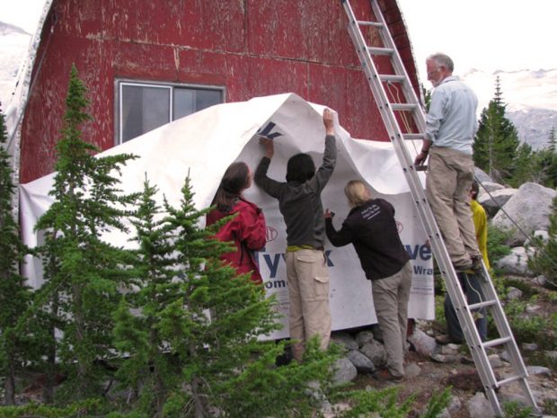 Tyvek, white plastic sheet, being applied over the weathered end-wall. Roland Burton stands on the silver ladder looking on at the 4 members holding up the Tyvek.