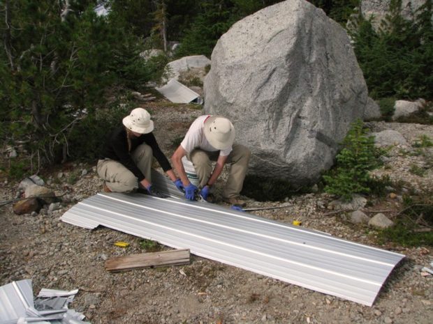 A short section of aluminum siding sits flat on the ground. A man using tin snips sits on a bucket is trimming of a section. A woman crouches next to the man holding the end being snipped and the larger piece of siding.