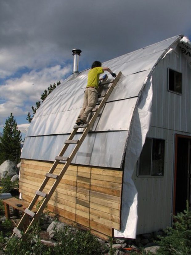 A man wearing a yellow shirt and tan pants is standing on the hand built wooden ladder screwing in a section of aluminum siding.