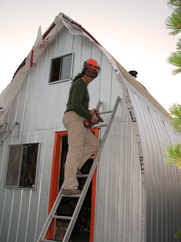 Portrait of a man wearing an orange helmet holding an electric drill while standing on an aluminum ladder near the front entrance to the Hut.