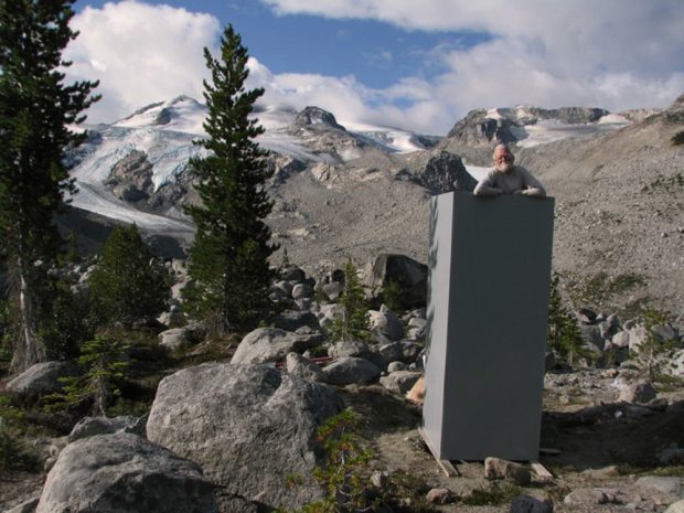 A man with a white beard and glasses rests his elbows against the roofless outhouse structure. Evergreen trees dot the boulder field behind and near the outhouse. A glacier laden peak can be seen in the distance.