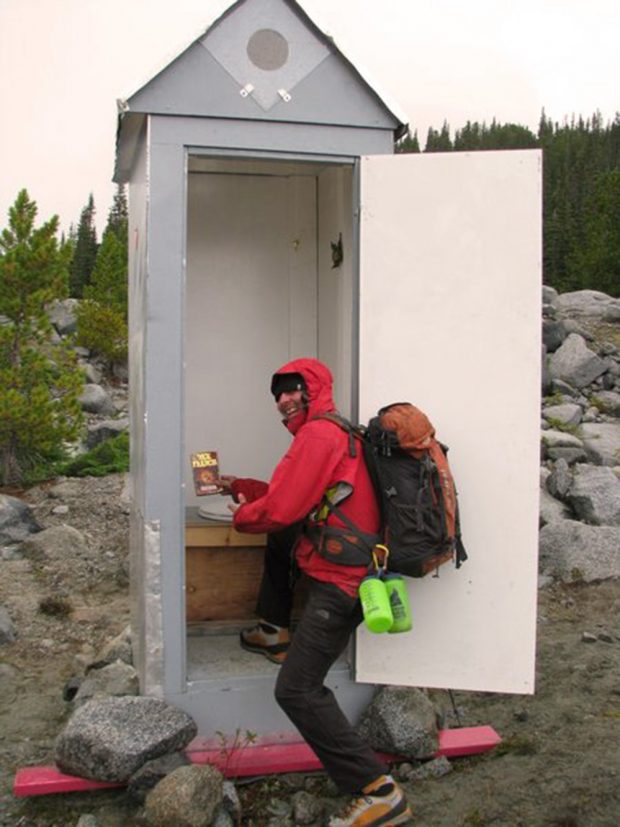 A man holding a paperback in his right hand, wearing a red jacket with the hood up and an orange and grey backpack poses with one foot inside the completed outhouse.
