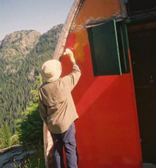 A man wearing a white hat is repainting the front end-wall of the Hut from a weathered orange to a bright red.