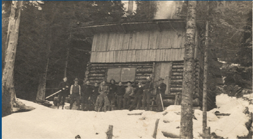 Black and white photograph, a Group of men stand in a line in front of the log cabin. Snow covers the ground around the cabin but the trees in the background are bare.