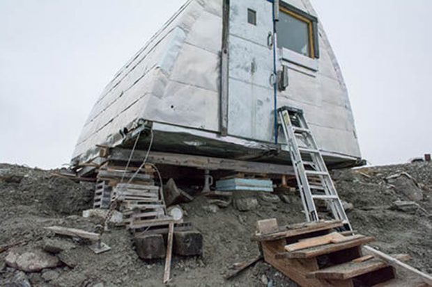 Aluminum siding wrapped Gothic arch hut supported by wood blocks because the earth below has been swept away. Wooden staircase can be seen in the foreground no longer attached to the hut.