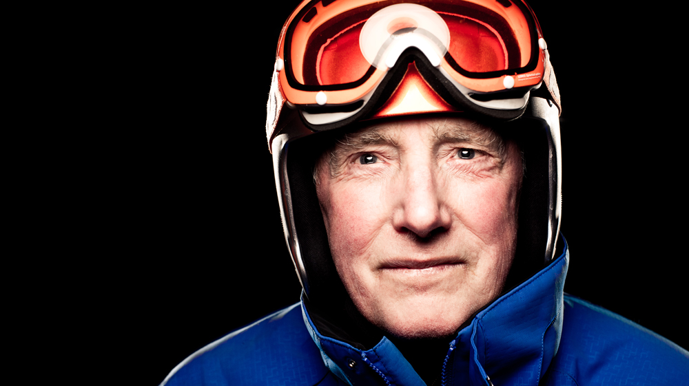 Close up of man wearing a ski helmet, ski goggles with orange lenses and a blue jacket and a black background.