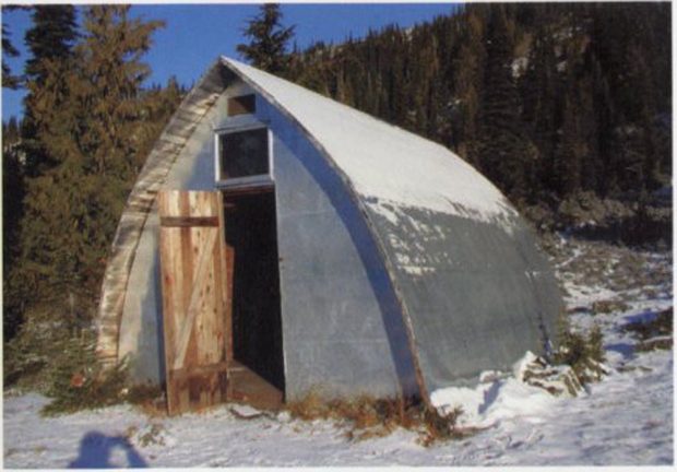 View of the front entrance to a Gothic arch Hut with the front door ajar. A light dusting of snow sits on the arched roof with evergreen trees in the background.