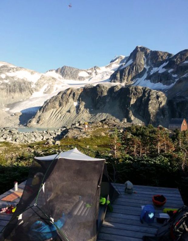 A black mesh tent sits on a wooden platform and across the green meadow dotted with trees sits the hut and in the background is the peak of Mount Wedge.