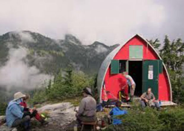 A group of hikers sit out front of the hut facing toward the hut entrance. One member is standing and another is sitting on the front stairway face towards the camera.