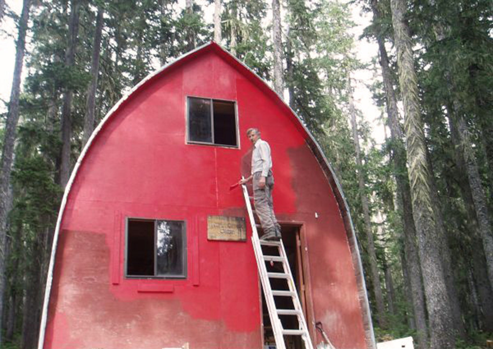 A man stands near the top of a retractable silver ladder holding a paintbrush and the painting is almost complete on the upper half of the Hut.