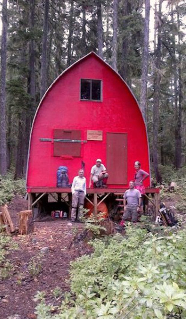 3 men and a woman stand outside the front entrance of the hut. The hut is a bright red and stands out from the light green leaves of the low-lying shrubs around the cabin. An old ski is placed across the shuttered window located next to the front door of the hut.