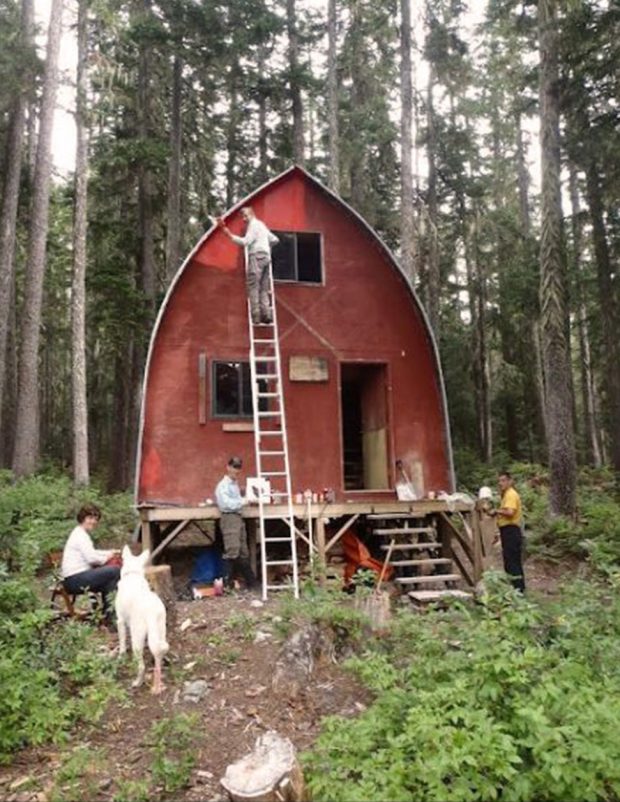 Club members have just begun repainting the Hut exterior. One man stands on an aluminum ladder and others pose around the front of the hut. A white haired dog is also visible.
