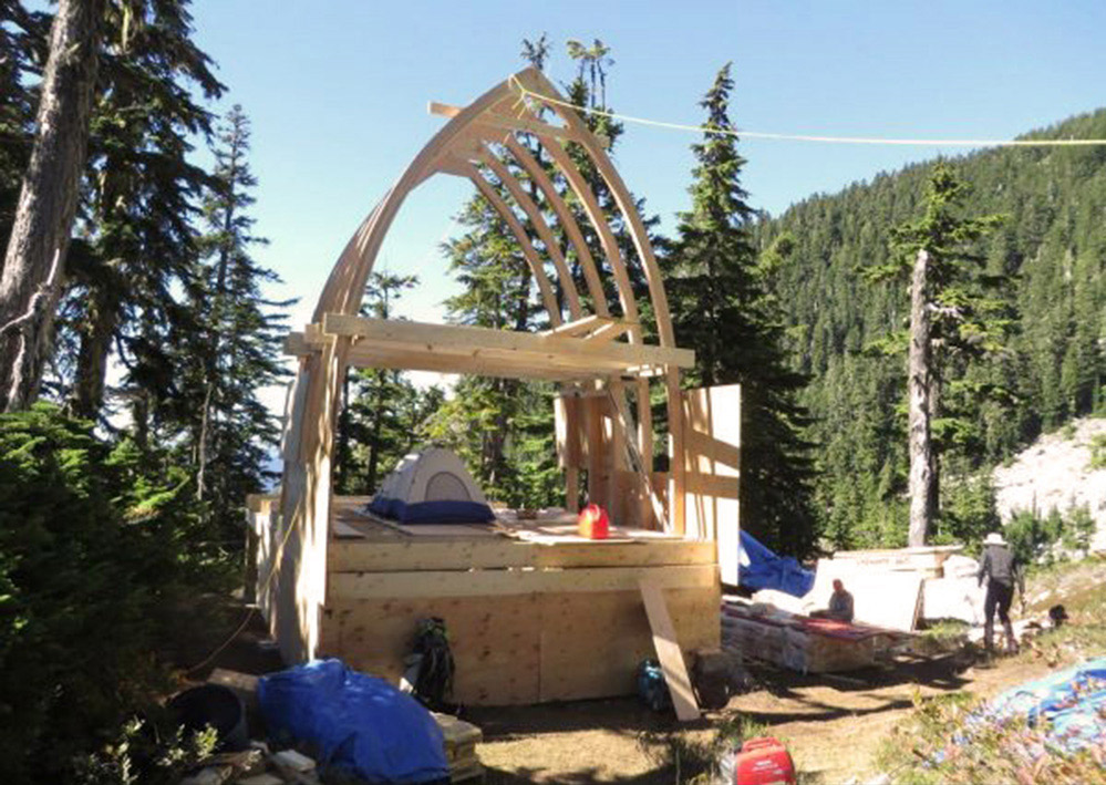 The wooden frame of the gothic arch hut during construction. A silver and blue tent sits on the main floor of the hut and a red gasoline can sits in front of it. Off to the right hand side, two workers can be seen standing near piles of lumber.