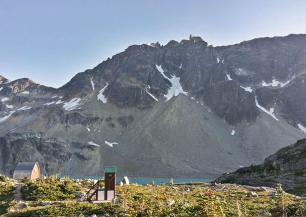 The Outhouse painted brown with a green roof stands in a meadow dotted with shrubs and small boulders and the Hut is on its left. Behind is the turquoise water of the lake and dark grey rock of the mountain peak.