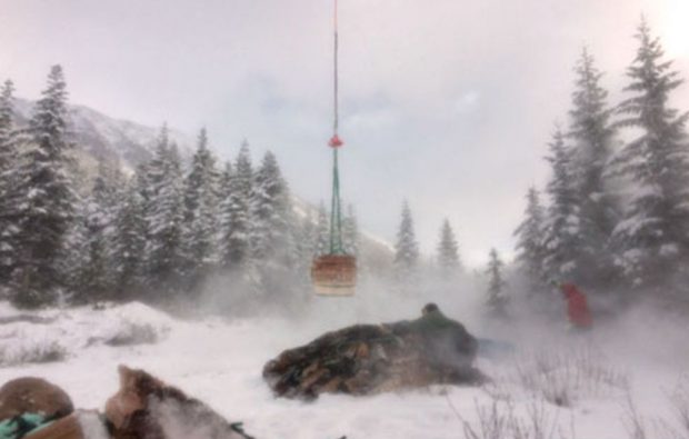 Long cable hangs holding supplies from a helicopter, which is not included in the photograph. One man is crouched behind a load of supplies and another stands off to the far right away from the blowing snow from the helicopter’s take-off.