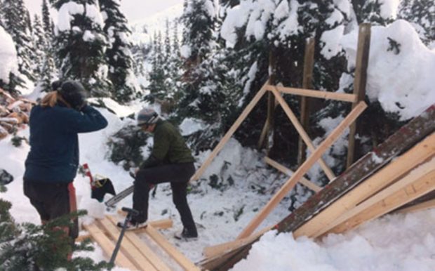 Rough framing for a new firewood shed has been erected and a member holding a chainsaw looks to trim two by fours on the ground while another member looks on.
