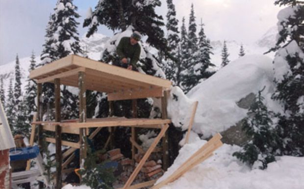 A man kneels on top of a newly framed structure attaching the roof to the firewood shed and deep snow surrounds the structure and covers the evergreen trees.