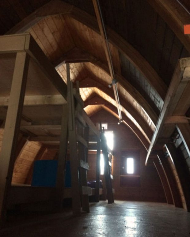 Sunlight shines in the windows of the far end wall and large wooden bunks line the left hand side of the second story sleeping quarters of the Hut.