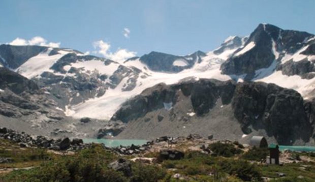 The summer sun beats down on the large glaciated peaks looming behind the blue-green water of the lake, which stands out in contrast to the dark grey rock. The Hut and outhouse is off to the right.