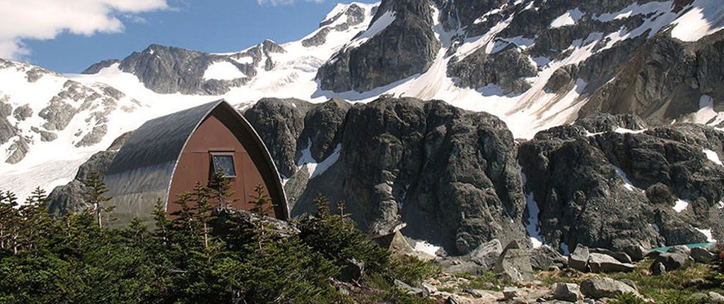 A Gothic arch hut surrounded by green evergreen trees sits in the sunshine with steep snow covered slopes in the background.