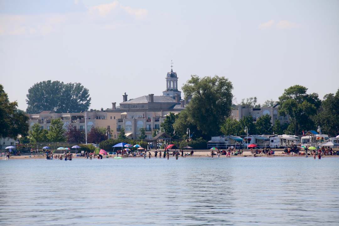 colour photograph from the water showing a beach with bathers in the water, multiple colourful umbrellas. To right are number of campers. In background is cupola of Victoria Hall