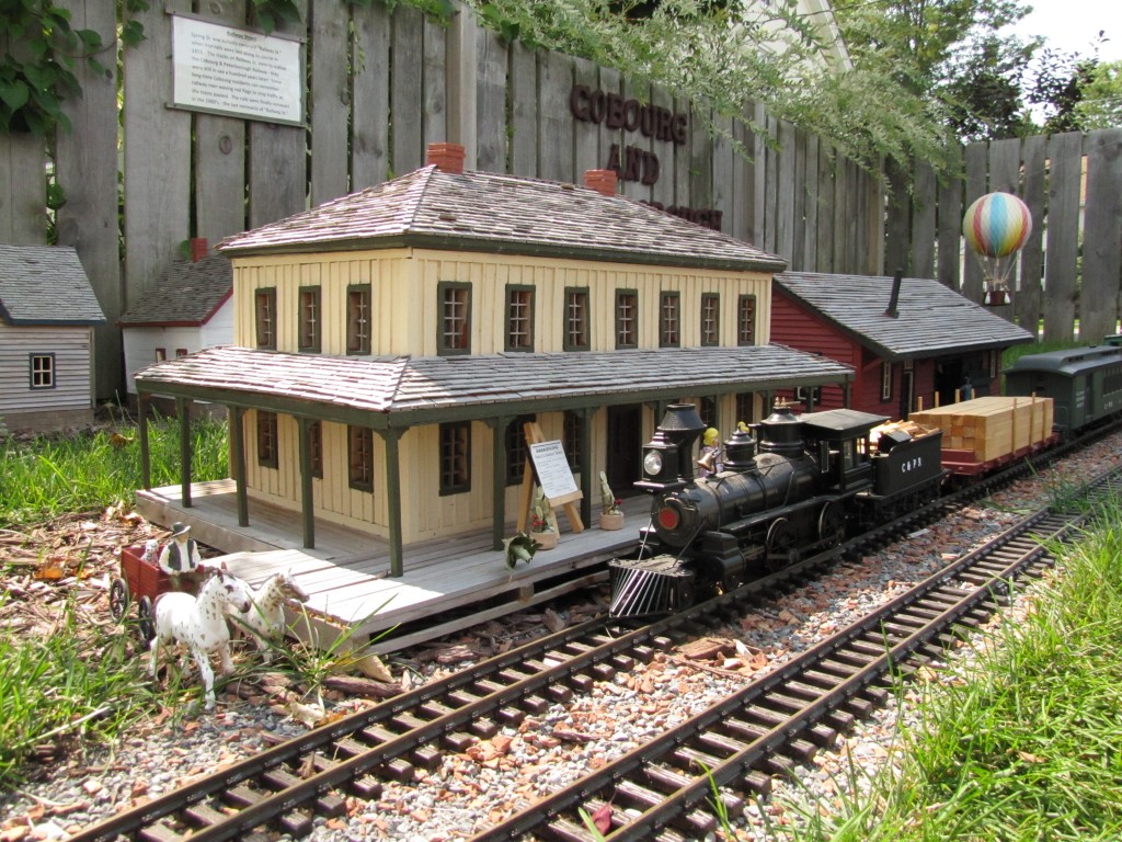 a colour photograph of 1/20 scale model of an antique black steam engine pulling a load of lumber in front of a two-story yellow station with a wood shake roof and a veranda. Two white horses with a cart and driver are near the tracks and a colourful hot air balloon flies in the background.