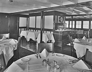 black & white photo of a sparse dining area with shiny wooden floor and carpet runners. Round tables are set with white table cloths, napkins, seven pieces of silverware and plates. Serving tables are in the middle background