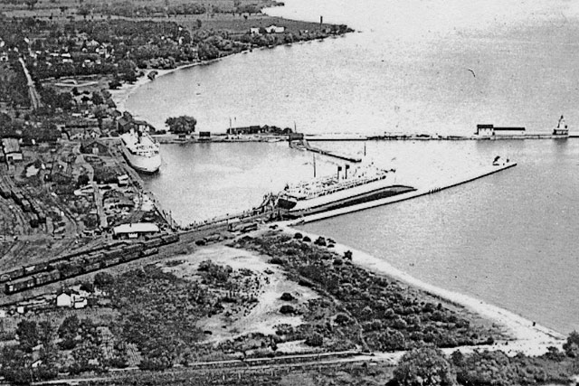 a black and white aerial photograph showing two ferries docked in Cobourg Harbour, one in the centre of the picture at a pier with its bow toward the lake and it's stern at the end of a railway line with railway cars on it, and the other at a dock along the shore. The picture includes the western extension of the harbour in the foreground and the beach in the distance.