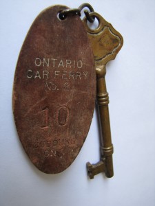 A bronze coloured skeleton type key attached by wire to a key fob made of brown material stamped with the text ONTARIO CAR FERRY No. 2 10 COBOURG ONT.