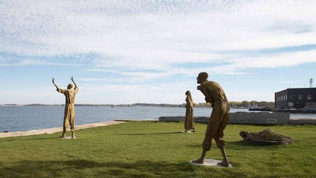 four bronze statues on a patch of green grass by the shore, standing at a distance from each other, turned towards the lake, one statue has its arms raised, one statue is a stooped, another is lying on the ground