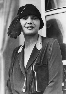 a black & white photo of a woman with full lips looking at the camera, with a purse under her left arm and a bowl shaped hat with large feather on her head