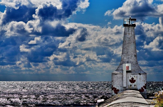 a colour photograph showing a blue sky with the sun reflecting off many clouds and dark water. In the right foreground is a round lighthouse with red maple leaves painted around the base.