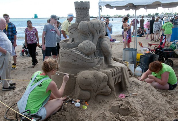 two female sand artists put the finishing touches to a large castle at blue water's edge, onlookers pause to admire the handiwork.