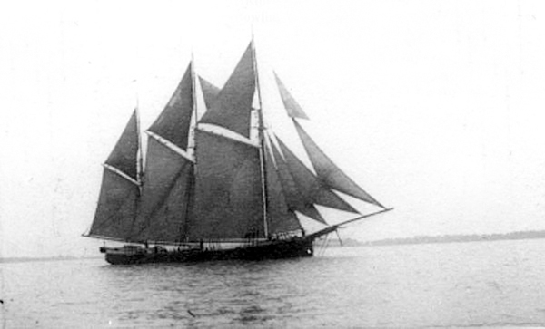 a black & white photograph of a sailing vessel with three masts and all sails up sailing by a distant shoreline