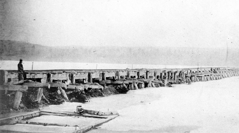 an old black and white photograph showing a rough wooden trestle bridge, nearby to the left and receding into the distance to the right over a lake which appears to be ice covered. A man sits on the bridge in the foreground and dimly in the distance is the other shore of the lake.