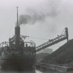 a dark black and white picture showing the stern view of a coal freighter on the left with a ramp carrying coal from the vessel and pouring it onto a huge coal pile to the right