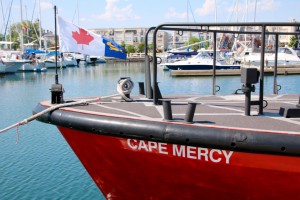 Photo en couleurs de la proue rouge du Cape Mercy faisant flotter le drapeau de la Garde côtière. Il y a des marins et des condominiums en arrière-plan.
