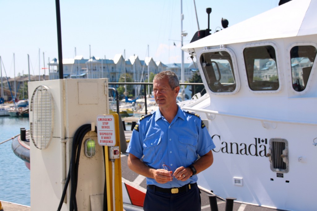 Photo en couleurs d’un homme habillé d’un uniforme d’été bleu. Il est debout devant un vaisseau sur lequel est écrit le mot « Canada ». Une grande pompe à essence se trouve à côté de lui.