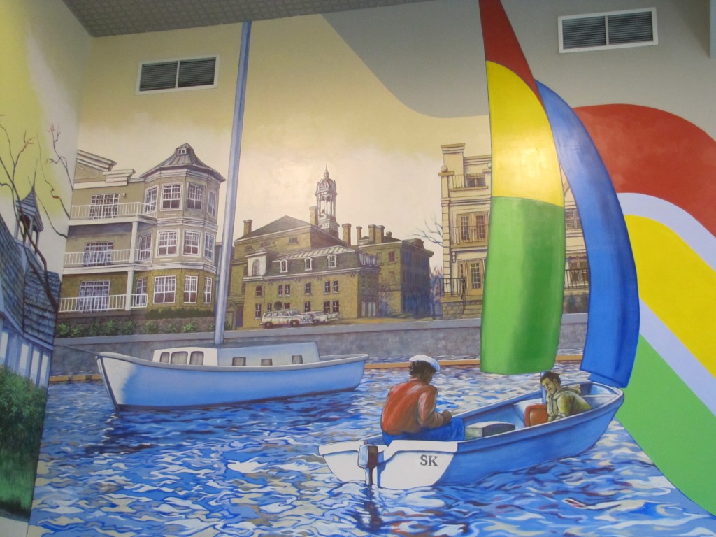 a colour photograph of a mural - in the foreground to the right, in blue slightly choppy water, is a small boat with motor, two sails and two passengers. It has the initials SK on the stern, one sail is red, yellow and green and the other blue. A second, larger sailboat, without sails raised, is to the left. On shore are brown and white condominium buildings with a space between through which can be seen brown Victoria Hall topped by its cupola clock tower. There are two air vents at the top of the mural