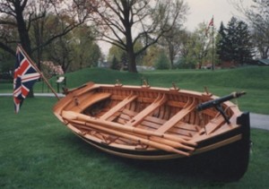 Petite embarcation de bois, avec un drapeau de l’Union Jack, repose légèrement inclinée sur l’herbe. Elle comprend quatre ensembles de dames et quatre bancs de rameurs, dont deux avec des trous pour les mâts.